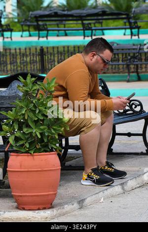 A man sits on a black wrought iron bench in the boulevard of Sancti Spíritus, Cuba, deeply focused on his smartphone. He is dressed in a warm yellow s Stock Photo