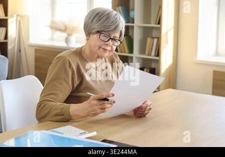 Senior woman reviewing paperwork and signing documents at home office table Stock Photo