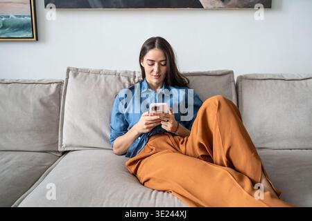 Tranquil woman painter resting on sofa scrolling media or texting online on smartphone at home. Stock Photo