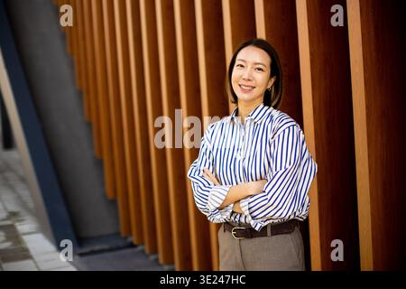 A woman stands with her arms crossed and smiles while leaning against a wooden wall. She wears a striped shirt and light-colored trousers. The place l Stock Photo