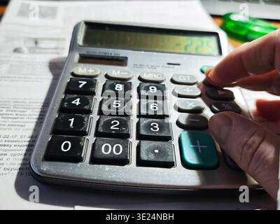 Hand interacts with a calculator displaying a number while papers are scattered on a desk in a work environment Stock Photo