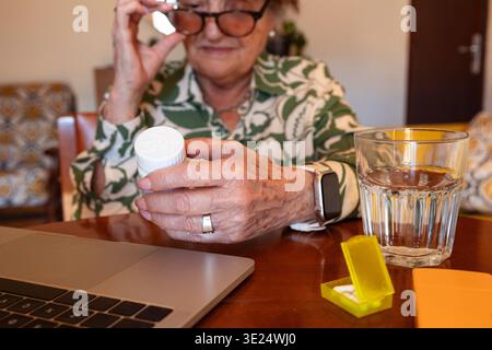 Senior woman reading prescription bottle, using a laptop, and preparing to take pills Stock Photo