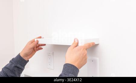 Mans hands holding white rectangular shelf plank against white wall near installed black bracket. Home improvement, positioning shelf board, DIY Stock Photo