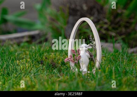 A white ceramic Easter bunny figurine holding pink and dried flowers stands in a large wooden oval frame on green grass, creating a charming festive Stock Photo