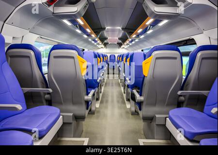 Interior view of a modern passenger train carriage featuring rows of ergonomic blue seats with yellow accents, a clean aisle, and bright LED lighting Stock Photo