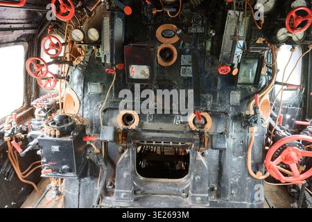 Detailed interior of an old steam locomotive control panel featuring numerous gauges, red valves, levers, and pipes in a historic train engine cabin. Stock Photo