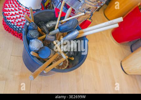 A bucket containing different types of percussion mallets and drumsticks placed on a wooden floor with colorful musical instruments nearby. Stock Photo