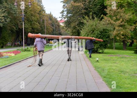 Three workers walk down a path in a park while carrying a long pipe. They are dressed in casual clothes and appear focused on their task. Stock Photo