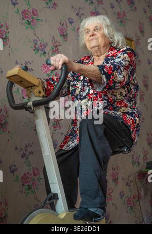 An elderly, gray-haired woman is exercising on a stationary bike at home. Stock Photo
