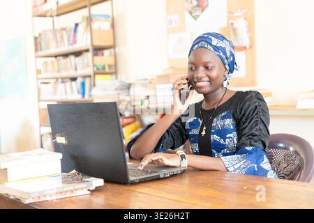 Beautiful African girl chatting on her phone while typing on her laptop Stock Photo