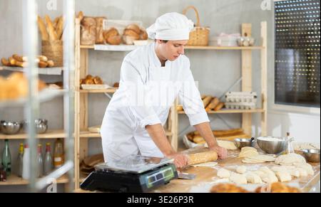 Young man baker in white uniform holding rolling pin in hands and rolling out dough in bakery kitchen Stock Photo