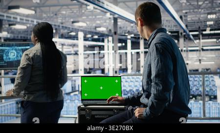 Data center IT experts investigate AI configurations using green screen laptop, making adjustments. Server hub engineers check deep learning errors on chroma key notebook, camera B Stock Photo