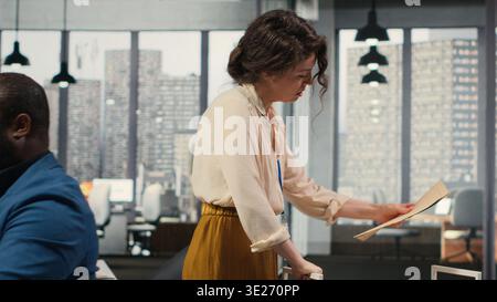 Workplace support employee moves a delivery cart stacked with folders and contracts, helping african american administrator bringing stack of archived documents for accounting tasks. Camera B. Stock Photo