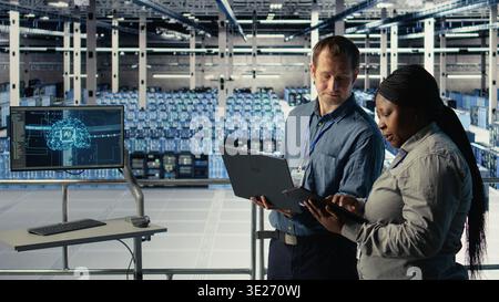 Server room coworkers use laptop to install updates and keep AI systems running. Data center engineering team uses notebook to write code improving infrastructure output, camera A Stock Photo