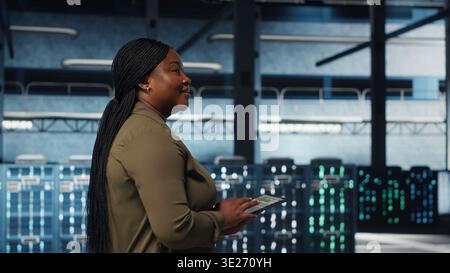 Smiling data center technician using tablet to manage software updates for system stability. Upbeat IT expert in server room configuring infrastructure with device for optimum performance, camera B Stock Photo