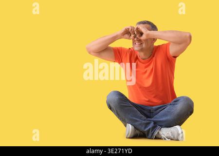 Mature man looking through imaginary binoculars on yellow background Stock Photo
