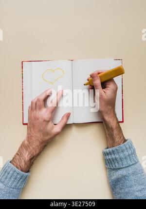 Male hands drawing a yellow heart with marker on open lined notebook. Neutral beige background, overhead view. Romantic concept of love, creativity an Stock Photo