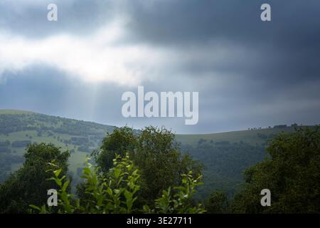Sun rays breaking through dark storm clouds over green hills. Atmospheric landscape with sun rays breaking through dark storm clouds above green hills Stock Photo