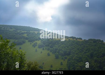 Sun rays breaking through dark storm clouds over green hills. Atmospheric landscape with sun rays breaking through dark storm clouds above green hills Stock Photo