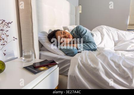 Asian woman in twenties lying on right side on bed, wearing light blue pajamas, holding smartphone Stock Photo