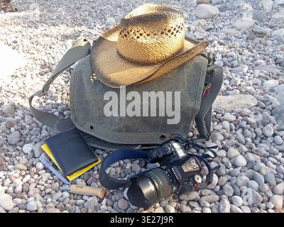 Photographer's outdoor gear arranged on pebble beach – camera, passport, Opinel knife, straw hat, and weathered canvas bag for travel and exploration Stock Photo