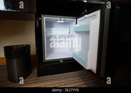 Empty mini fridge open in hotel room with carpet floor and trash bin nearby, typical hotel amenity Stock Photo