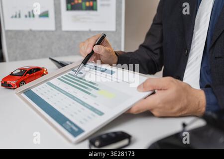 A businessman in a suit is calculating loan details, reviewing financial documents, and holding car keys near a calculator and toy car models, represe Stock Photo