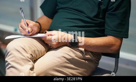 Person writing on paper indoors, focus on hands and pen, casual attire, engaged in note-taking or completing a task. Stock Photo