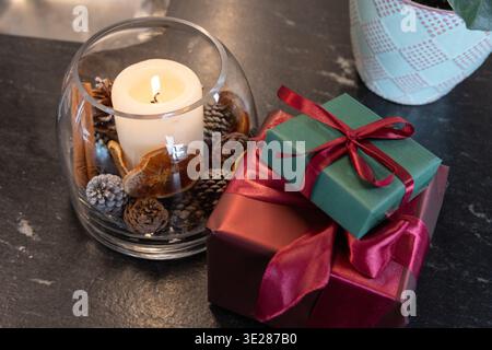 Glass bowl holding lit pillar candle, potpourri on slate tabletop, burgundy and green gifts sitting Stock Photo