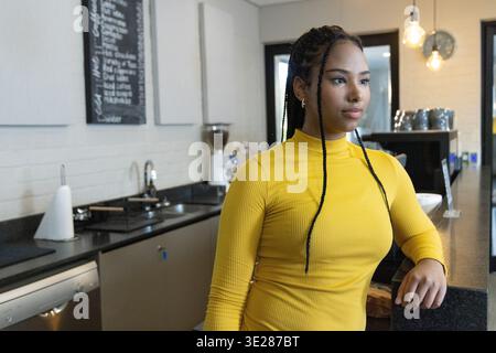 African American woman standing at cafe counter wearing yellow top, gazing right near espresso unit Stock Photo