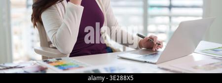 Professional female engaged in design development on laptop with pen in hand. Bright workspace with color palette nearby showing dedication productivi Stock Photo