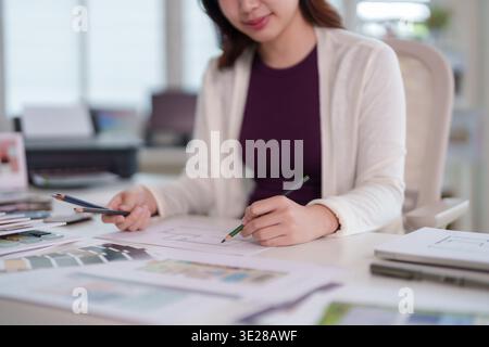 Professional Asian adult female designer diligently sketches new architectural plans using colored pencils in a bright modern workspace Stock Photo