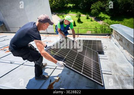 Workers building solar panel system on metal rooftop of house. Two men installers carrying photovoltaic solar module outdoors. Alternative, green and renewable energy generation concept. Stock Photo