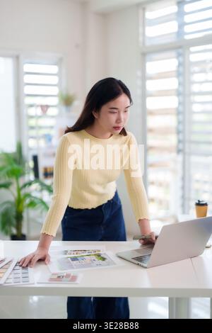Focused Asian female professional working on design projects remotely. She analyzes blueprints selecting materials for creative development. Stock Photo