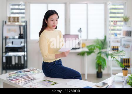 Asian adult woman reviews architectural plans and color swatches in her bright modern studio. She focuses on design project details and new ideas. Stock Photo