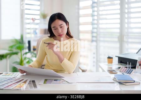 Asian adult woman designer meticulously analyzing architectural plans and color samples at a sunlit contemporary desk. Stock Photo