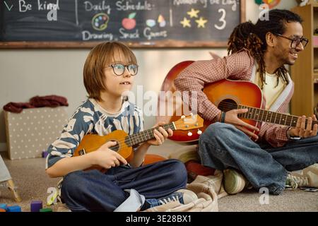 Boy Playing Ukulele with Black Man Teaching Guitar in Kindergarten Stock Photo