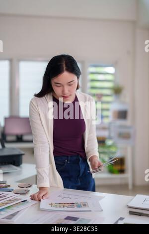 Asian adult professional woman reviews blueprints material swatches at her design studio. She focuses on architectural planning selecting colors textu Stock Photo