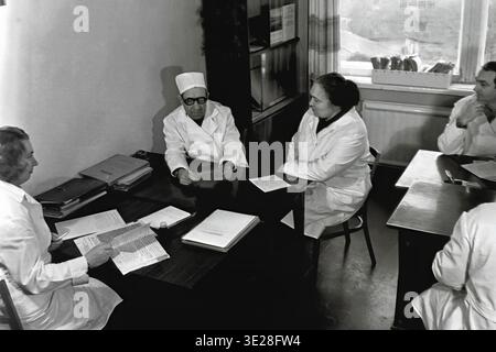 Vintage black and white photograph from 1982 capturing a medical briefing at the AIZ Hospital in Slavyansk. A group of four Soviet doctors in white coats is seated around a wooden desk in an office, carefully reviewing patient records and discussing clinical cases. The desk is covered with diagnostic paperwork and medical charts. This authentic historical archival image beautifully reflects the collaborative teamwork and occupational routine of healthcare professionals, preserving the era of a peaceful Donbas. Stock Photo