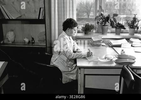 Vintage black and white photograph from 1982 providing a wider view of a female medical professional in a white coat working at a desk inside an office at the AIZ Hospital in Slavyansk. She is filling out medical records next to a window lined with potted plants and a wooden bookcase. This authentic historical archival image beautifully illustrates the working environment, administrative clinical routine, and occupational paperwork of Soviet medicine, preserving the era and the everyday life of a peaceful Donbas. Stock Photo