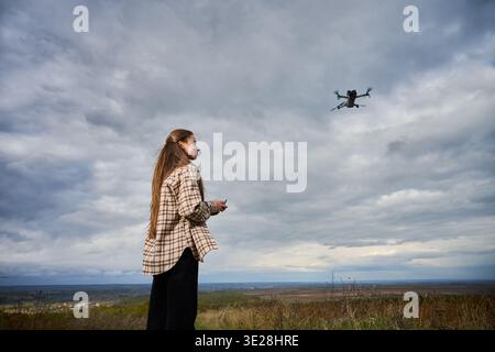 Woman piloting drone in cloudy sky over open landscape. Side view of female operator with controller on hill, aerial photography and exploration concept of technology Stock Photo