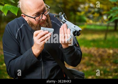 Man inspects drone while holding battery before installation. Bearded man checking quadcopter details in park, preparing equipment for flight and ensuring proper setup, concept of modern technology and maintenance Stock Photo