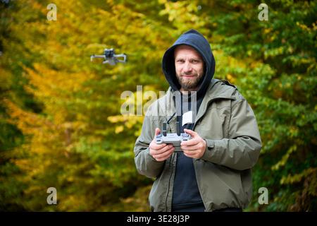 Man flying drone in autumn park, controlling quadcopter with remote controller while it hovers behind him, outdoor hobby and modern technology concept Stock Photo