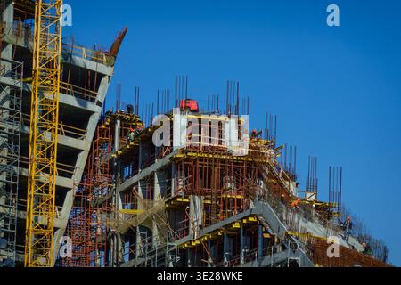 High rise building under construction, concrete skeleton with scaffolding and crane, construction workers in helmets, clear blue sky. Stock Photo