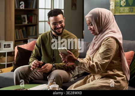 Young Muslim man smiling while holding tea cup, sitting beside young adult Muslim woman wearing hijab showing smartphone screen, both sitting on sofa indoors Stock Photo