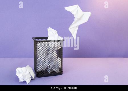 Origami bird flying from a waste basket filled with crumpled paper on purple background Stock Photo