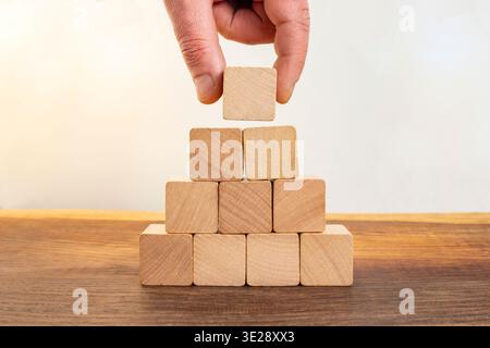 Hand placing final wooden block stacked pyramid structure built wooden cubes, for strategic growth, establishing strong foundation Stock Photo