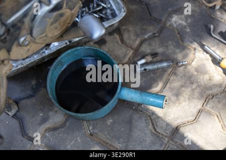 Used dark motor oil collected in blue plastic drain pan sitting on concrete paving blocks near various motorcycle parts tools and spark plug ready for Stock Photo