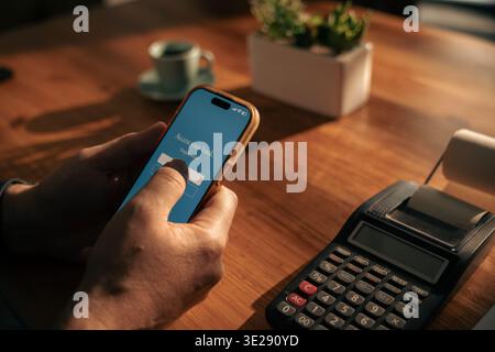 Mature man using smartphone for mobile banking and digital finance management at home with calculator on wooden desk. Stock Photo