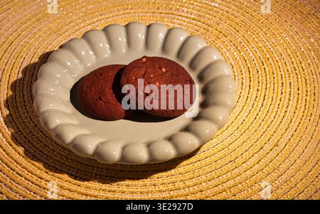 Two chocolate cookies on a scalloped ceramic plate, placed on a yellow textured placemat. Warm sunlight with long shadows, close-up shot Stock Photo